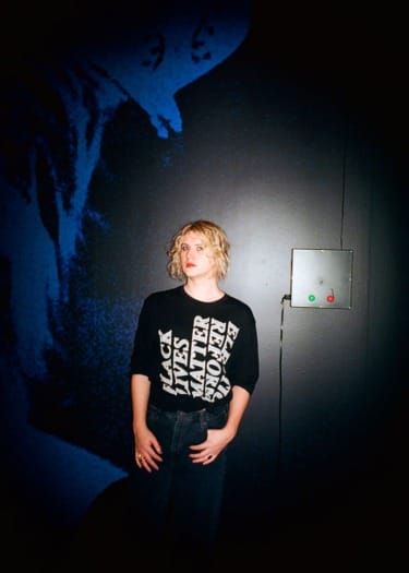 Person with curly hair wearing a "Black Lives Matter" shirt standing in front of a dark wall with blue artwork