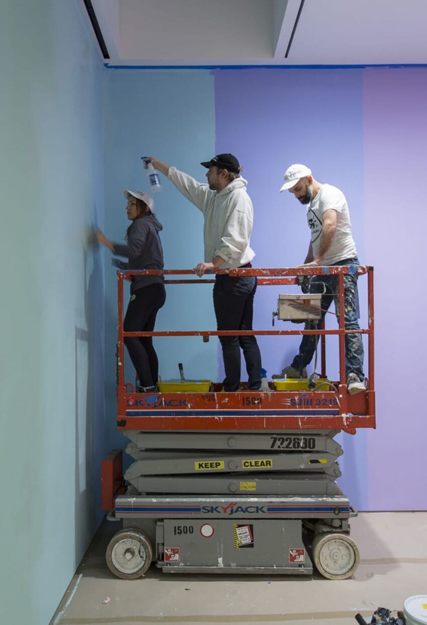 Three people on a lift painting a wall with blue and purple gradient in an art installation setting.
