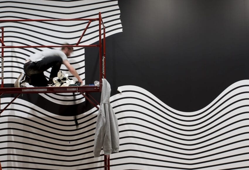 Artist painting wavy black and white lines on a large wall while standing on scaffolding, with a hoodie hanging nearby.
