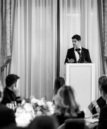 Man in suit speaking at podium in formal setting with seated audience in foreground, black and white image.