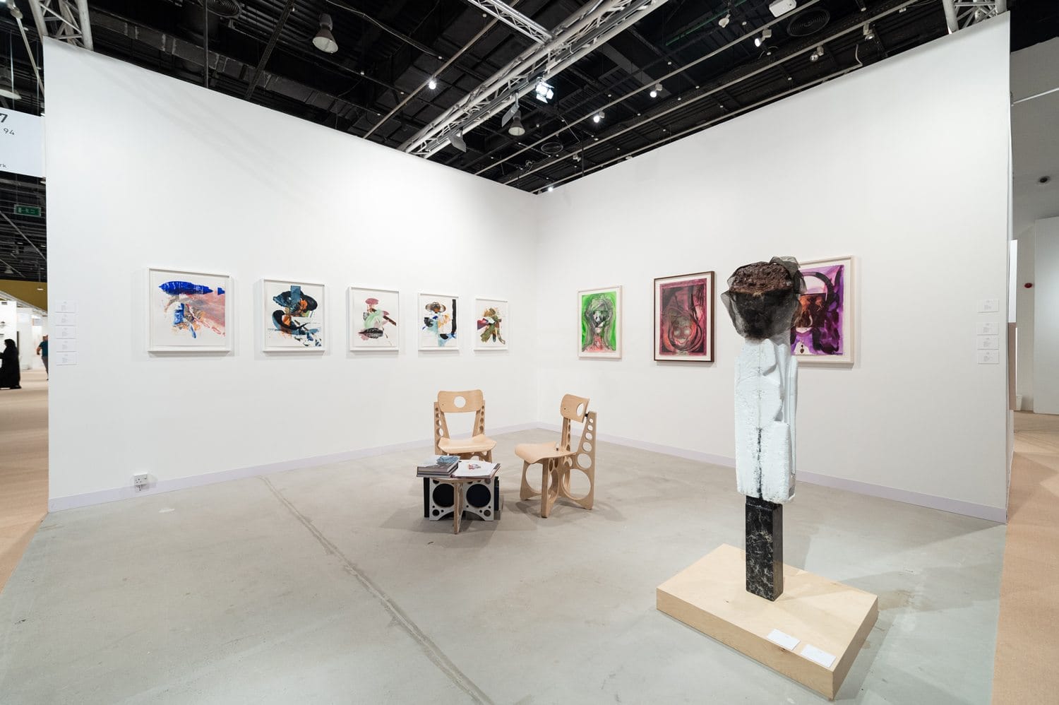 Art gallery interior with white walls displaying colorful paintings and a modern sculpture, wooden chairs in the center.