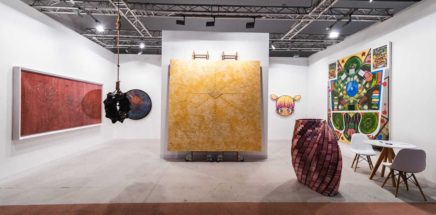 Art gallery with diverse contemporary paintings and sculptures displayed under industrial-style ceiling lights.