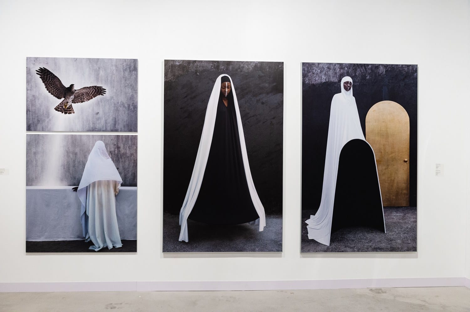Art gallery display featuring three photographs of figures in black and white cloaks with a bird image on the left