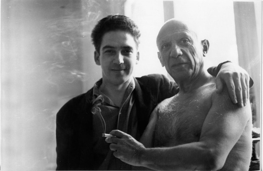 Two men in a black and white photo, one with a cigarette and the other shirtless, standing closely with arms around each other.