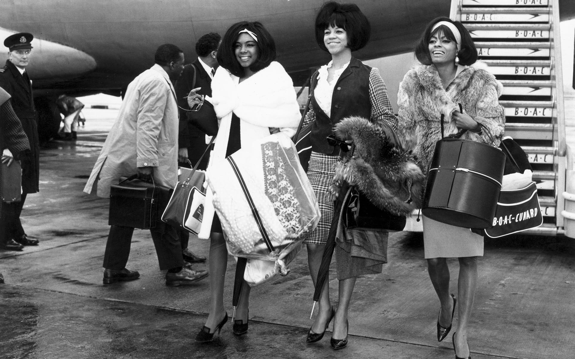 Three stylish women in coats holding bags and luggage, walking by an airplane on a tarmac, smiling and chatting.