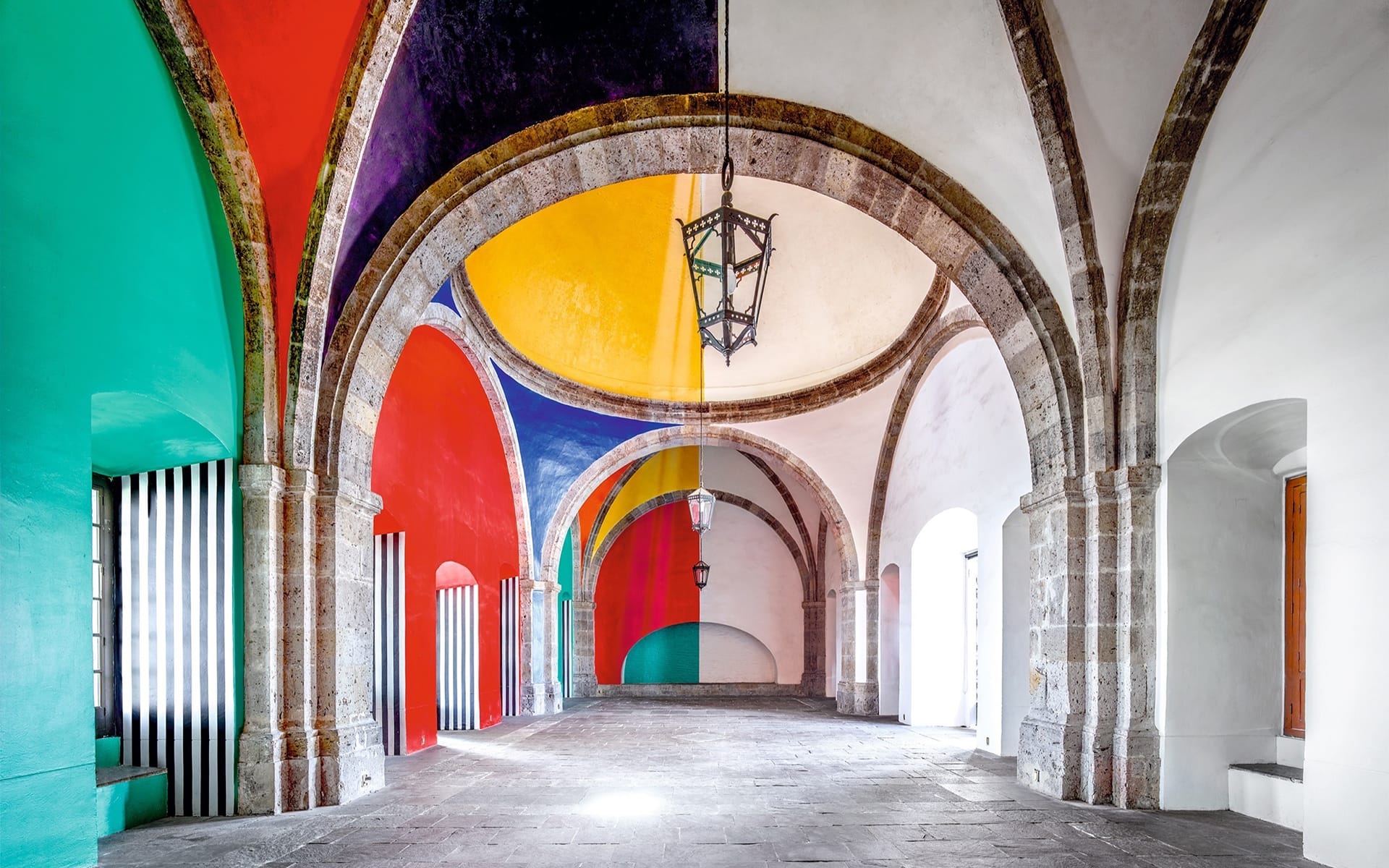 Colorful arched hallway with vibrant geometric patterns on walls and ceiling, featuring stone floor and hanging lantern.