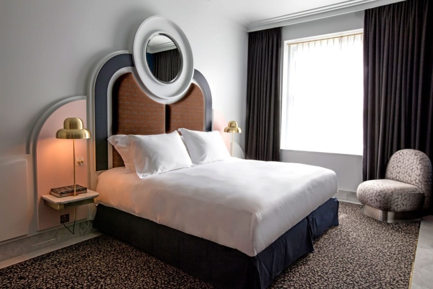 Modern hotel room with a white bed, stylish headboard, two bedside lamps, and a large window with dark curtains.