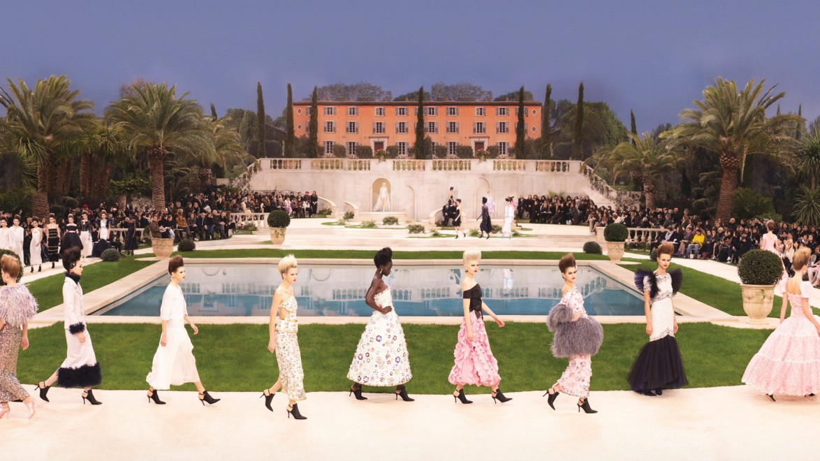 Fashion show with models walking in elegant outfits around a pool, outdoor setting with spectators and lush greenery.