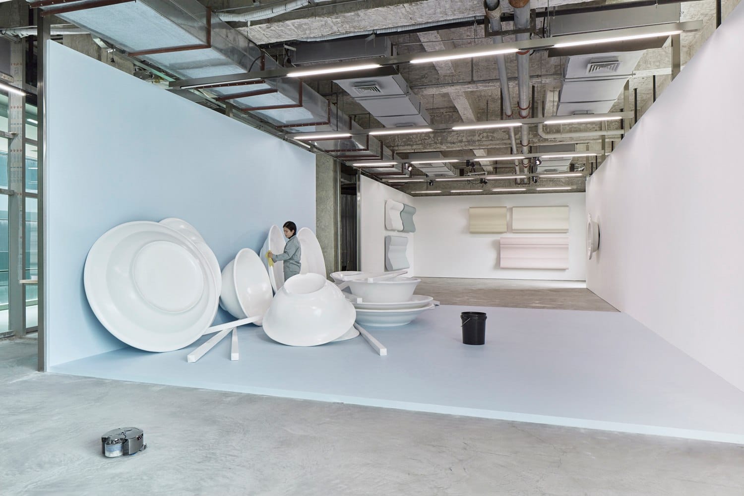 Person installing large white abstract sculptures in an industrial-style gallery space with concrete floors and ceilings.