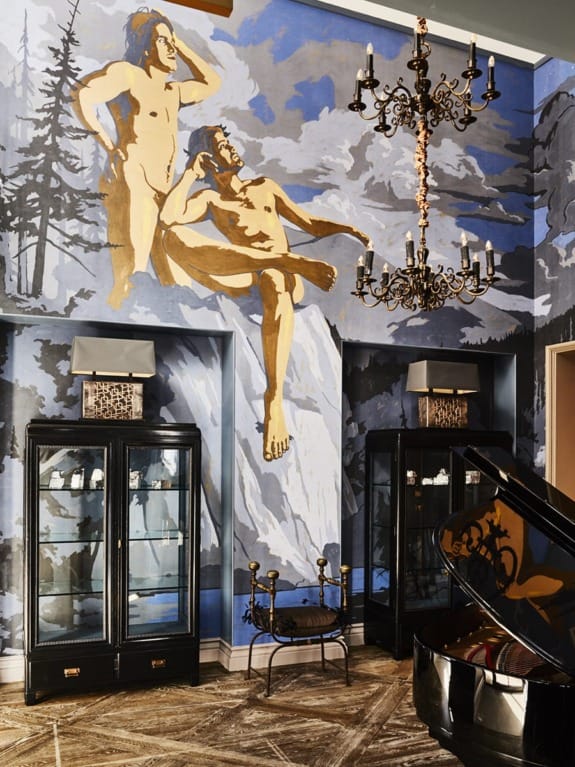 Interior with wall mural of two figures in nature, ornate chandelier, black display cabinets, and grand piano on patterned floor.