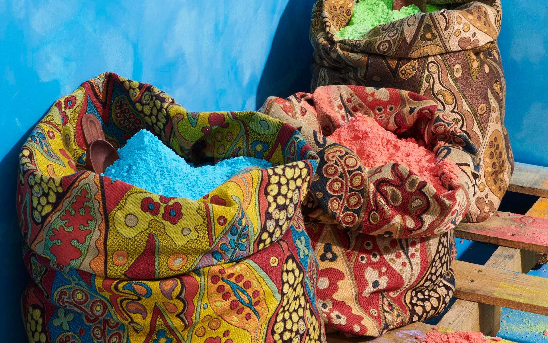Colorful patterned bags filled with vibrant blue, red, and green powders against a blue background on wooden pallets.