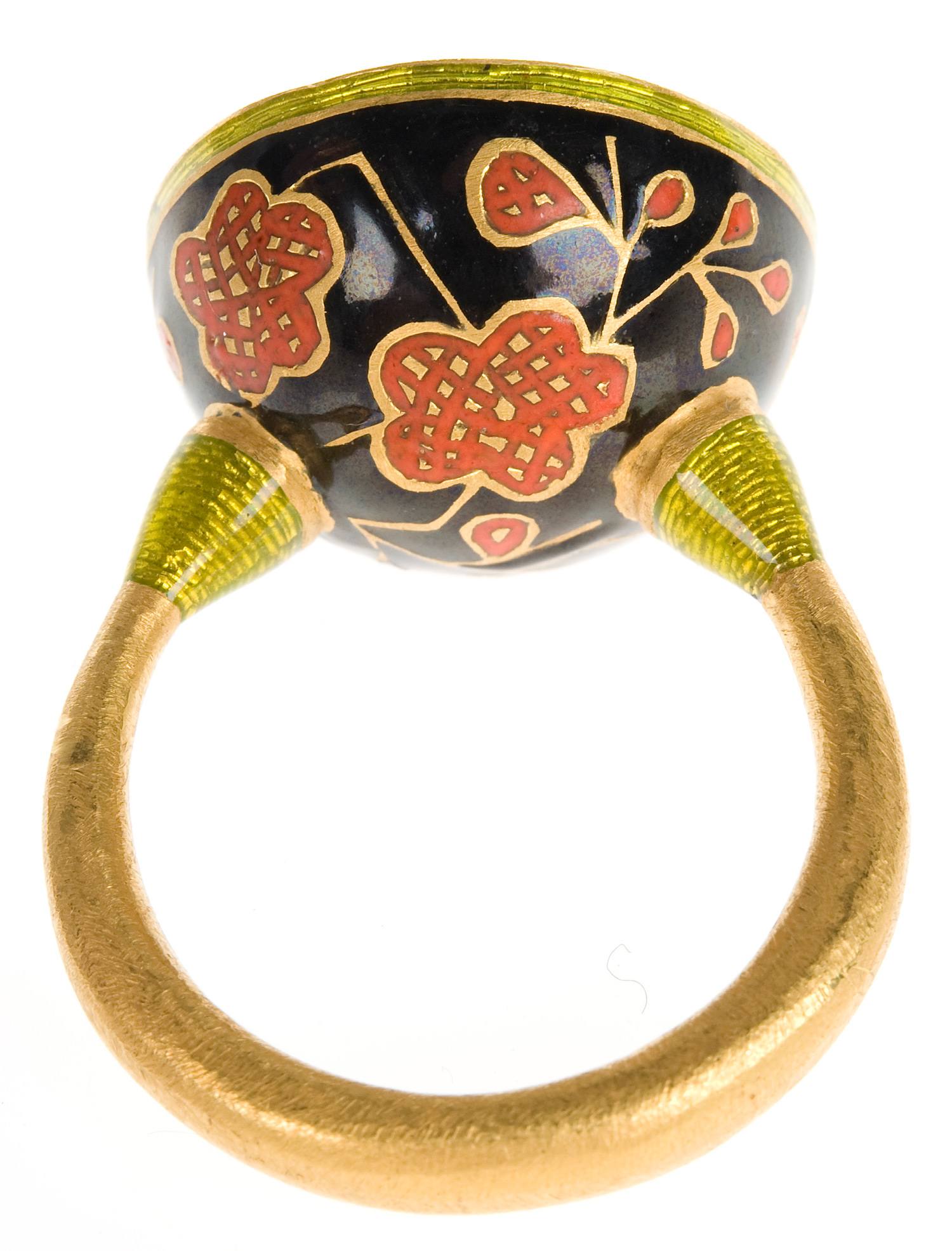 Gold ring with black and red enamel floral design on a round bezel, viewed from the side showing the decorative details.