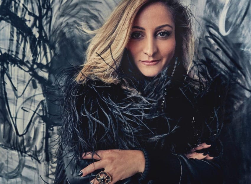 Person with long hair wearing a feathered outfit, standing in front of abstract art background.