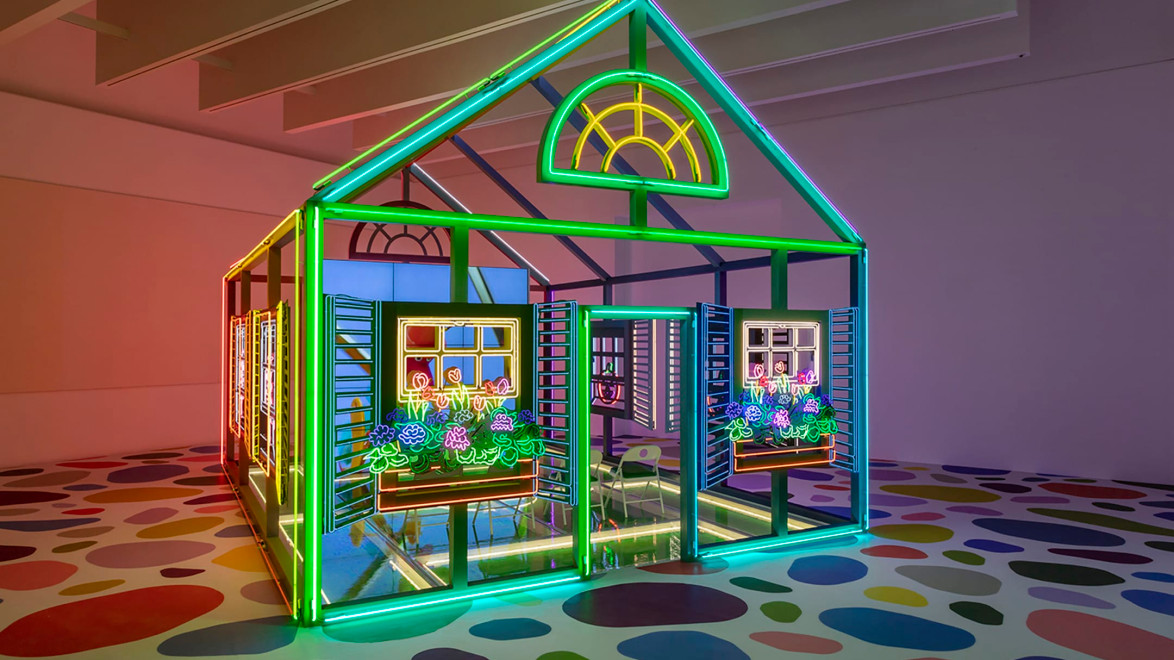 A colorful neon art installation shaped like a house with vivid window flower boxes, set on a polka-dot patterned floor.