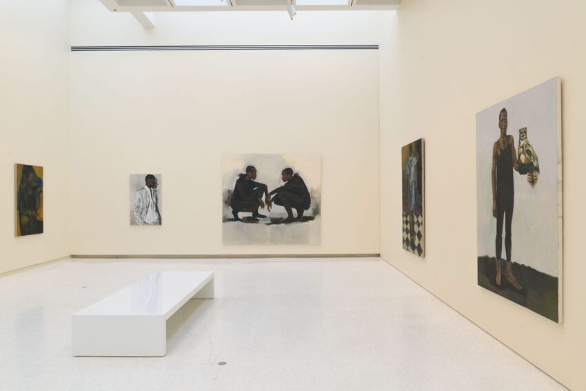 Art gallery with various modern paintings on white walls and a white bench in the center of the room.