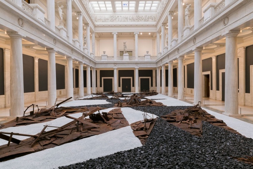 Contemporary art installation in a grand hall with marble columns and skylight, featuring abstract rusted metal and black gravel elements.