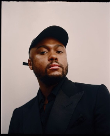 Person wearing a black cap and a dark suit looking confidently at the camera against a neutral background.