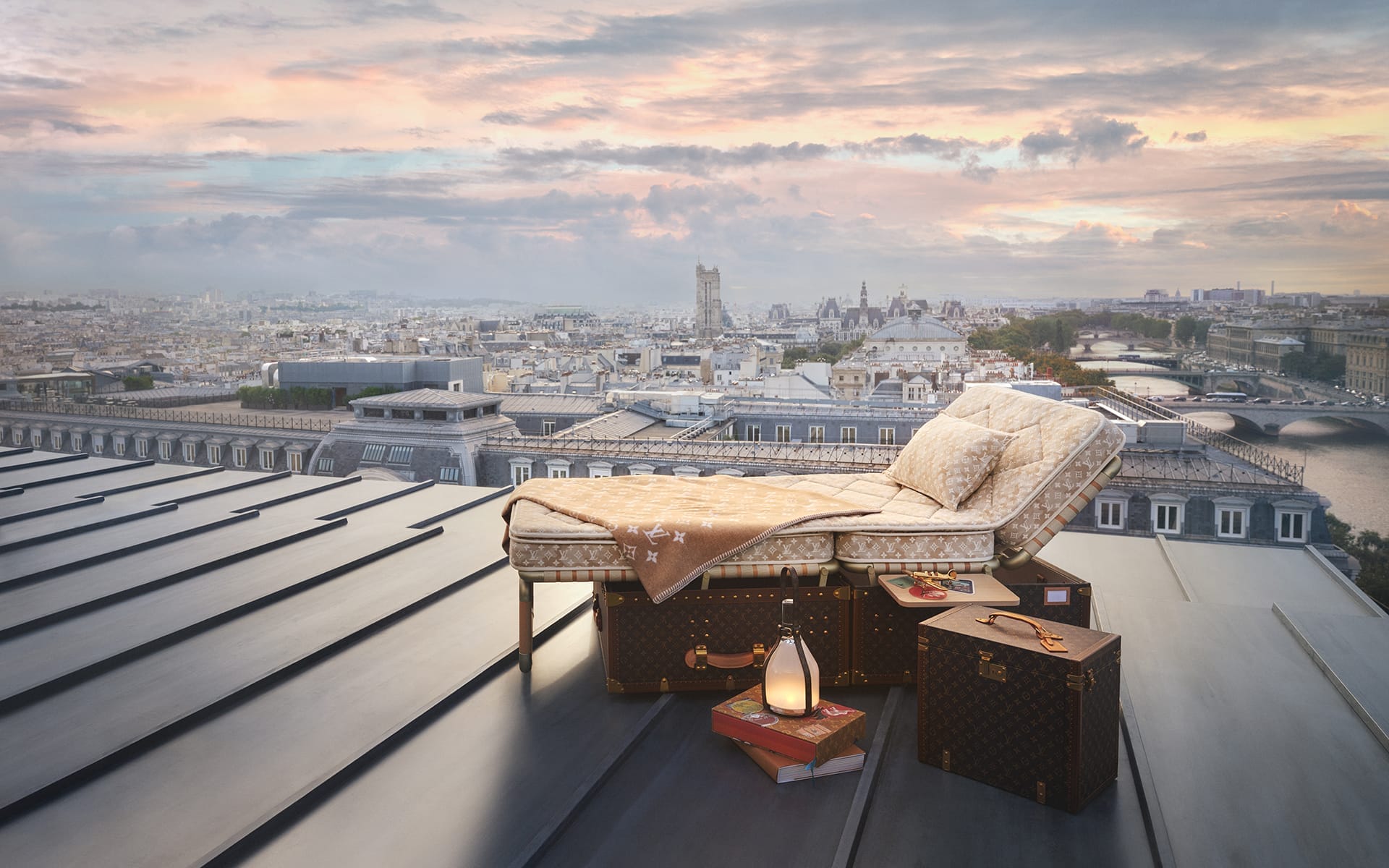 Outdoor bed setup on a rooftop overlooking a city skyline at sunrise with luxury travel trunks and a lantern nearby