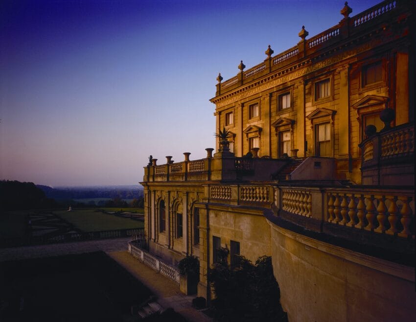 Historic mansion at sunset with ornate architecture and lush gardens, overlooking a vast landscape in the background.