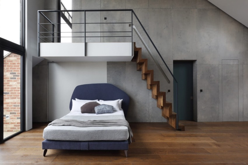 Modern bedroom with loft, wooden stairs, and a bed with blue headboard against a concrete wall and large window.