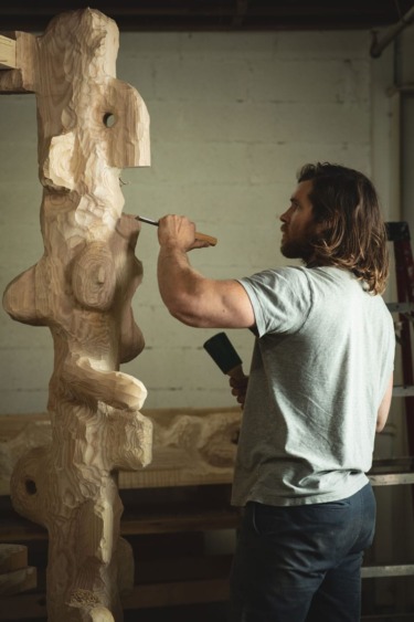 Artist carving a large abstract wooden sculpture in a studio space with focused expression and tools in hand.