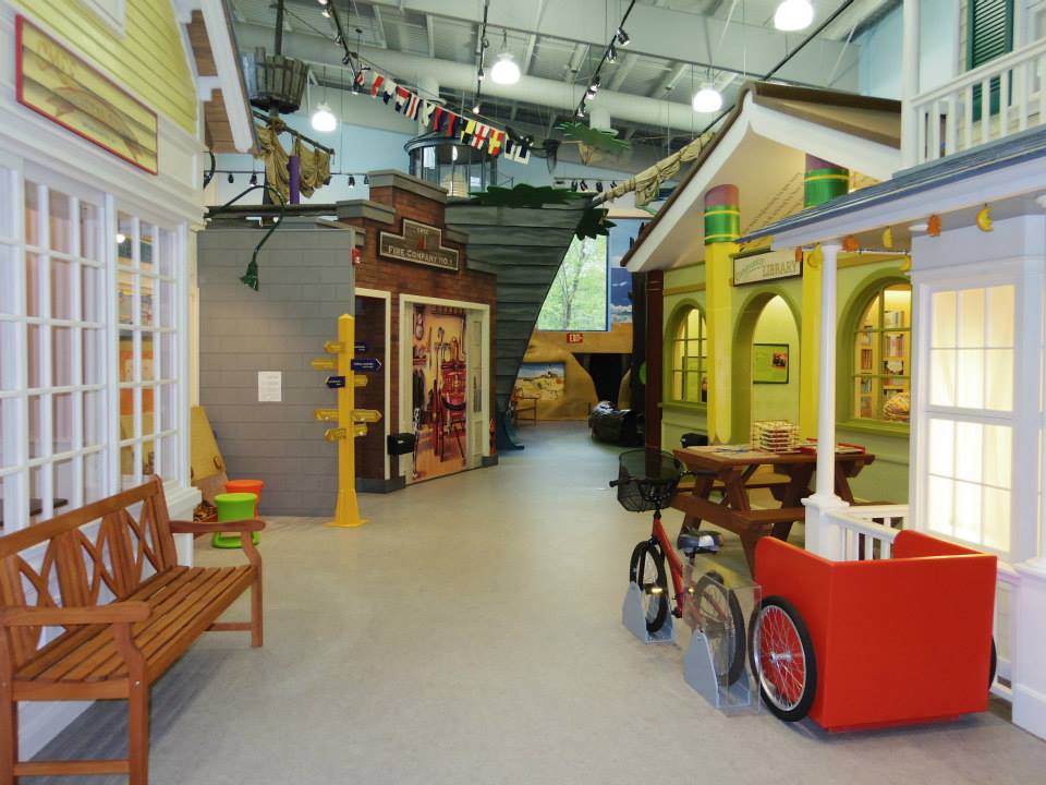 Children's museum village with colorful shops, play areas, and vibrant decor under a bright, spacious ceiling.