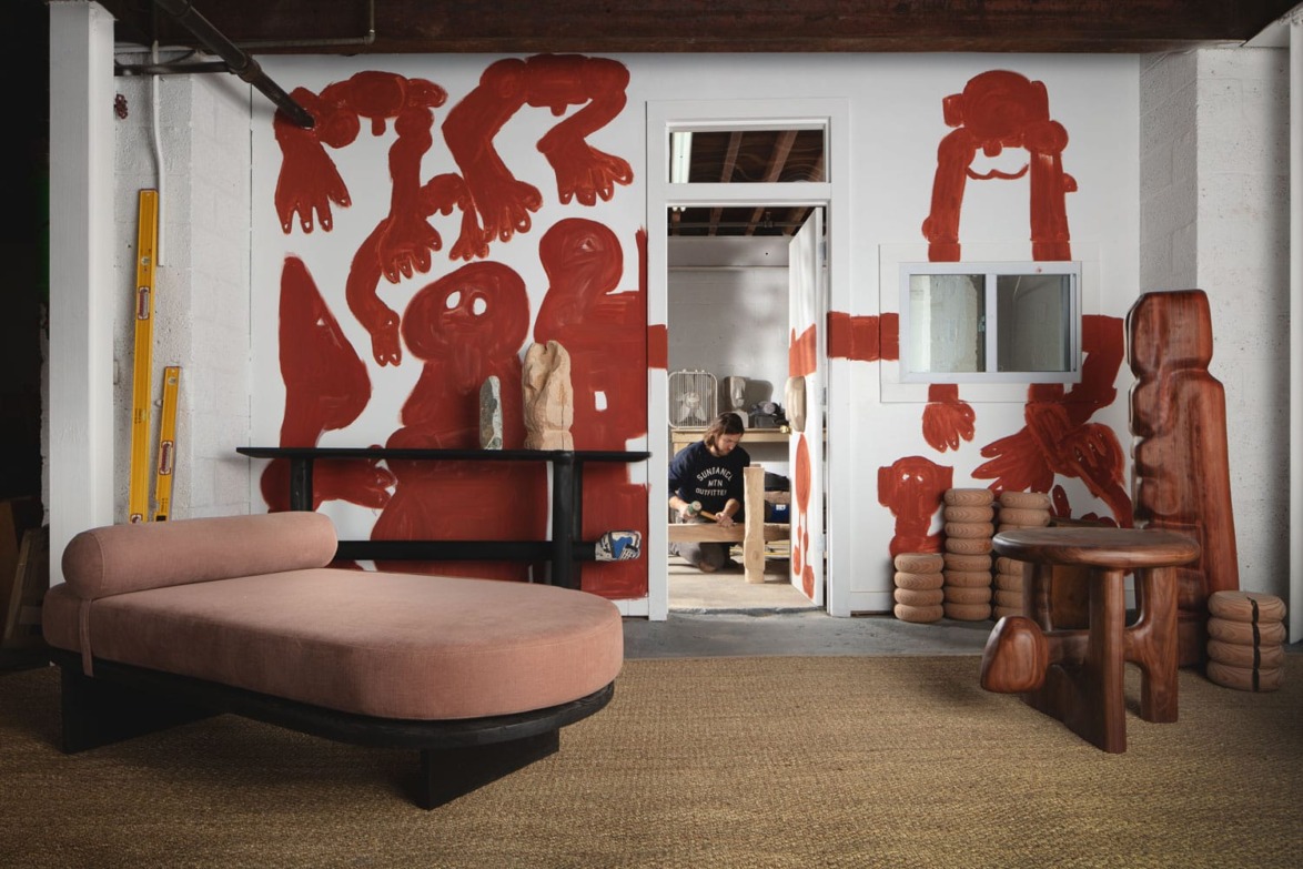 Artistic room with red humanoid wall murals, modern wooden furniture, a cushioned chaise lounge, and a person visible through the doorway.