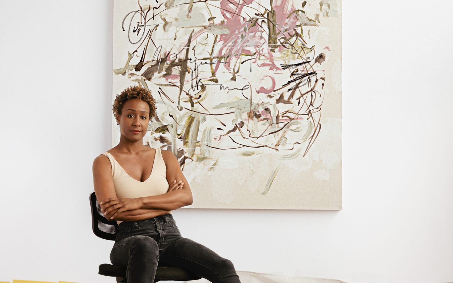 Person sitting on a chair in front of an abstract painting with arms crossed, wearing a light top and dark pants.