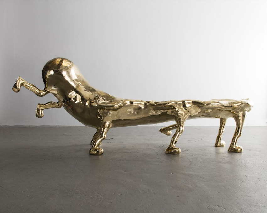 Sculptural artwork of a smooth, elongated golden creature with multiple legs on a gray floor against a white backdrop.