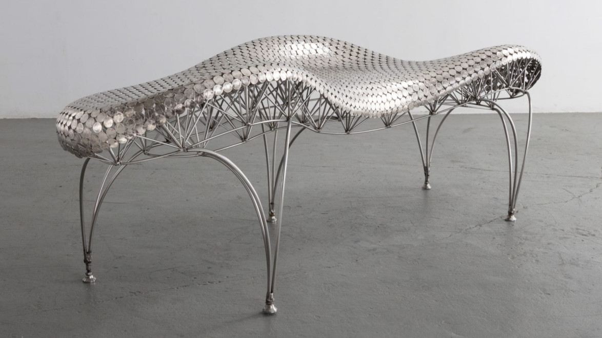 Futuristic metal bench with a wavy, textured surface and arching legs on a plain concrete floor.