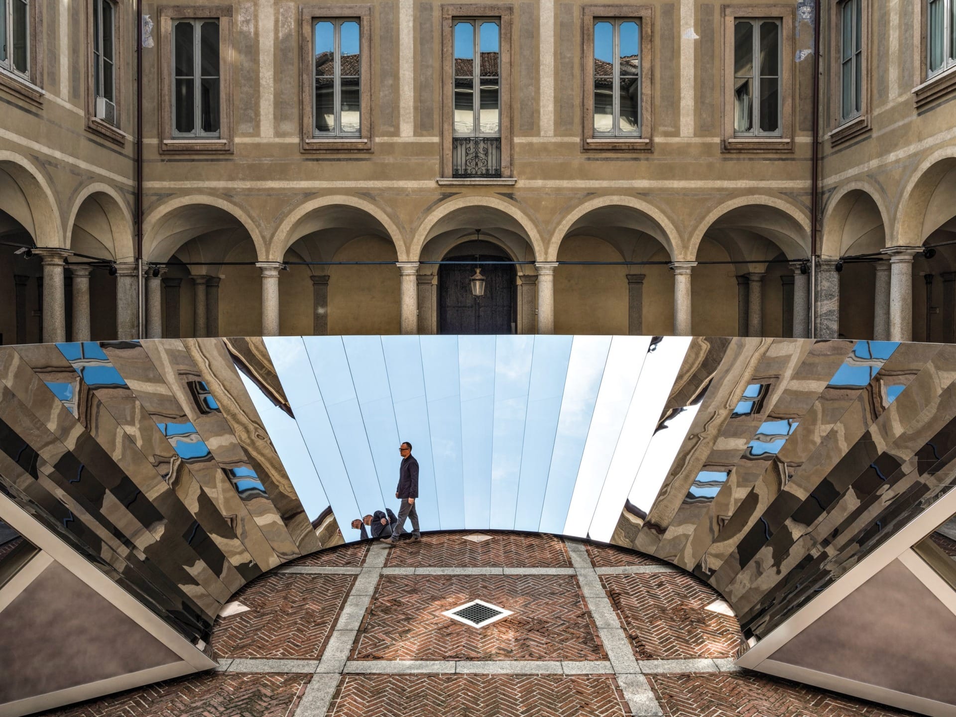 Person walking on mirrored structure in courtyard of historic building with arched windows and columns.
