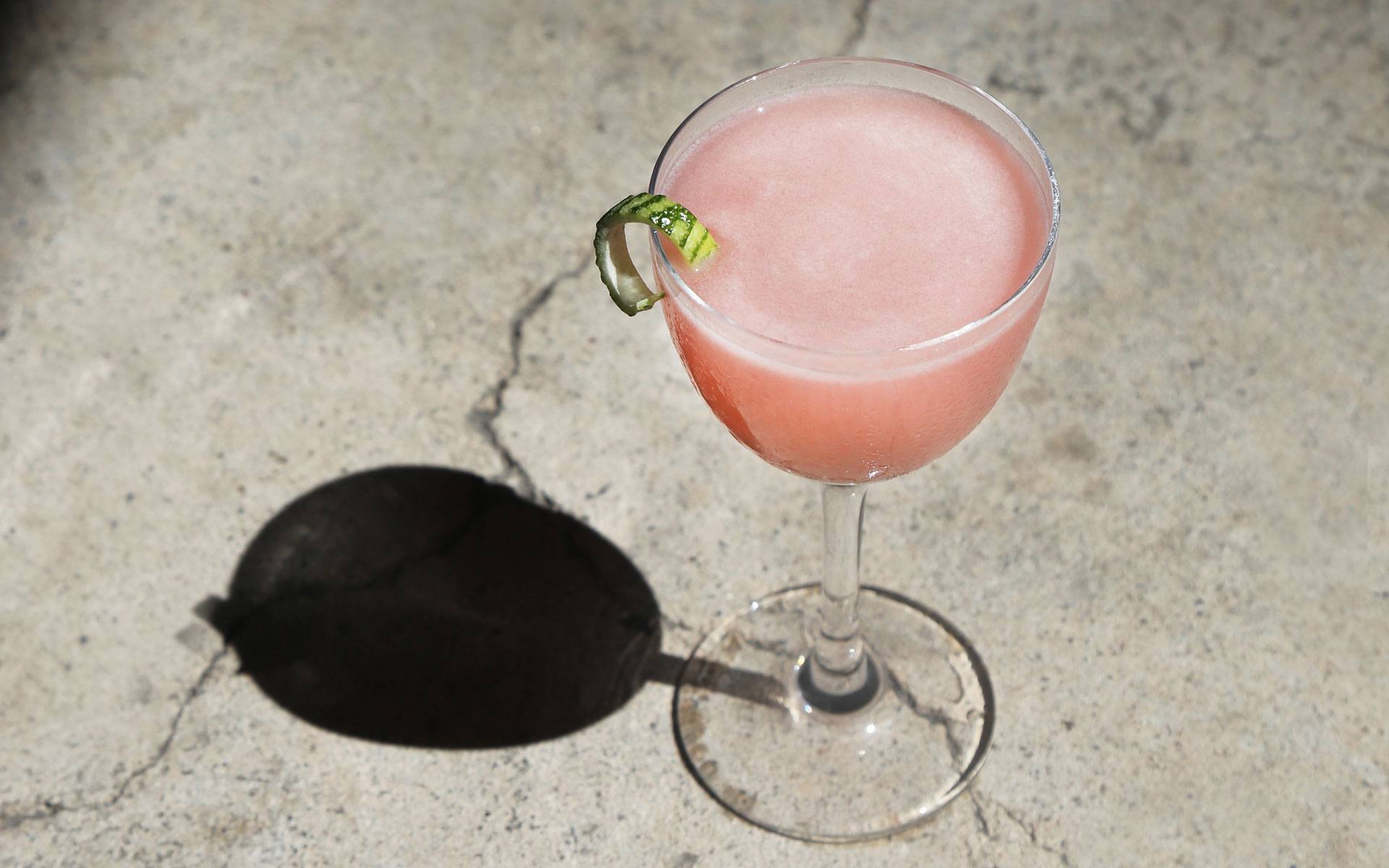 Pink cocktail in a stemmed glass with a lime twist garnish, casting a shadow on a concrete surface.
