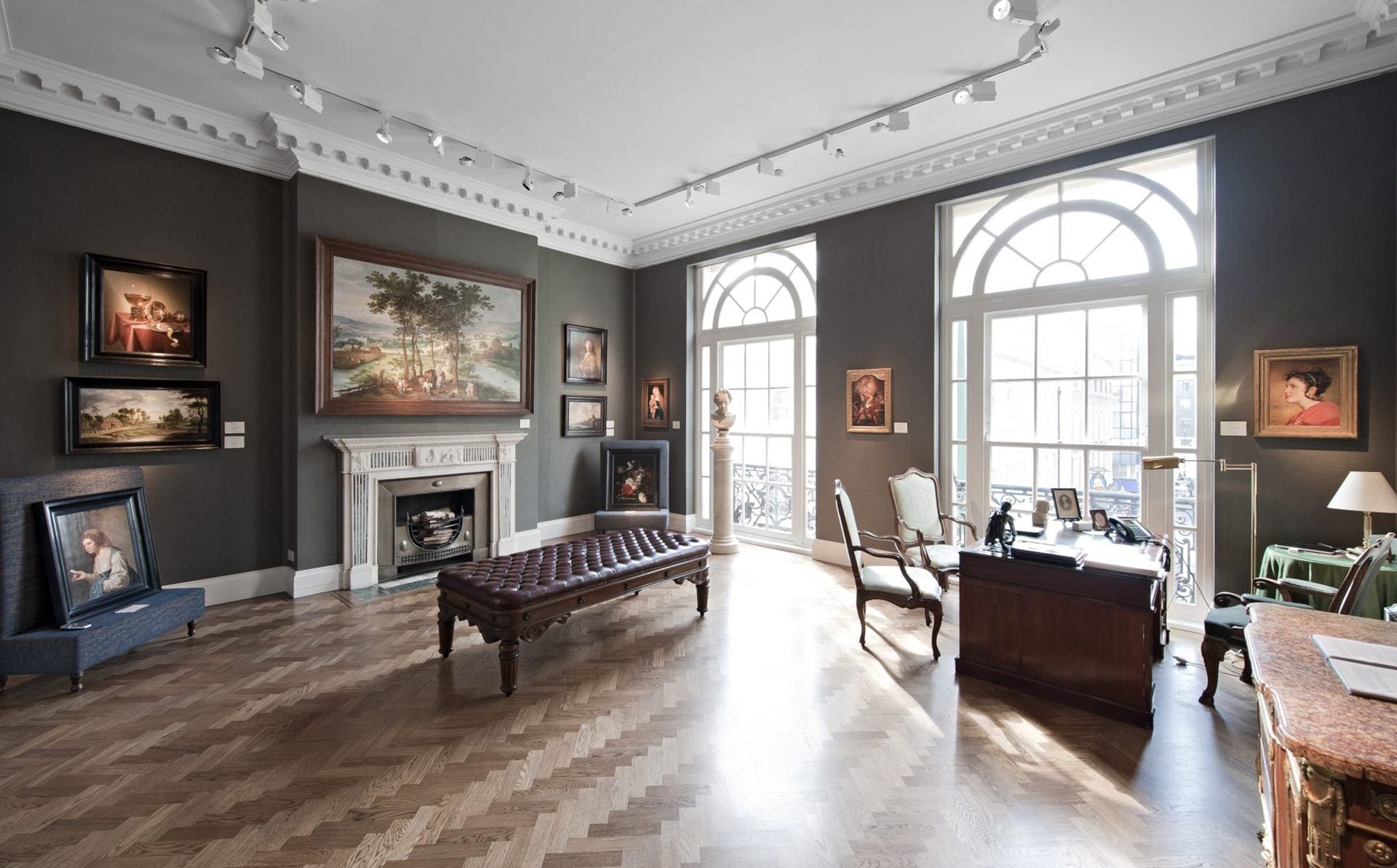 Elegant art gallery room with paintings on charcoal-colored walls, large windows, wooden floor, and classic furniture.