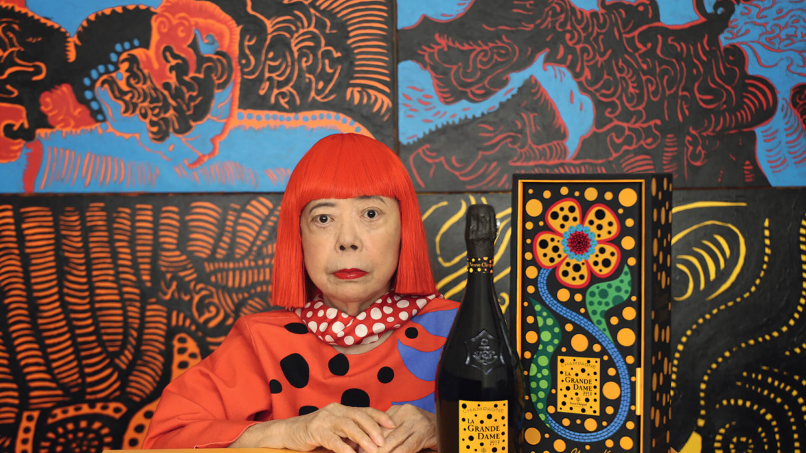 Artist in red wig and polka dot outfit next to a designed champagne bottle and box with a colorful abstract background.