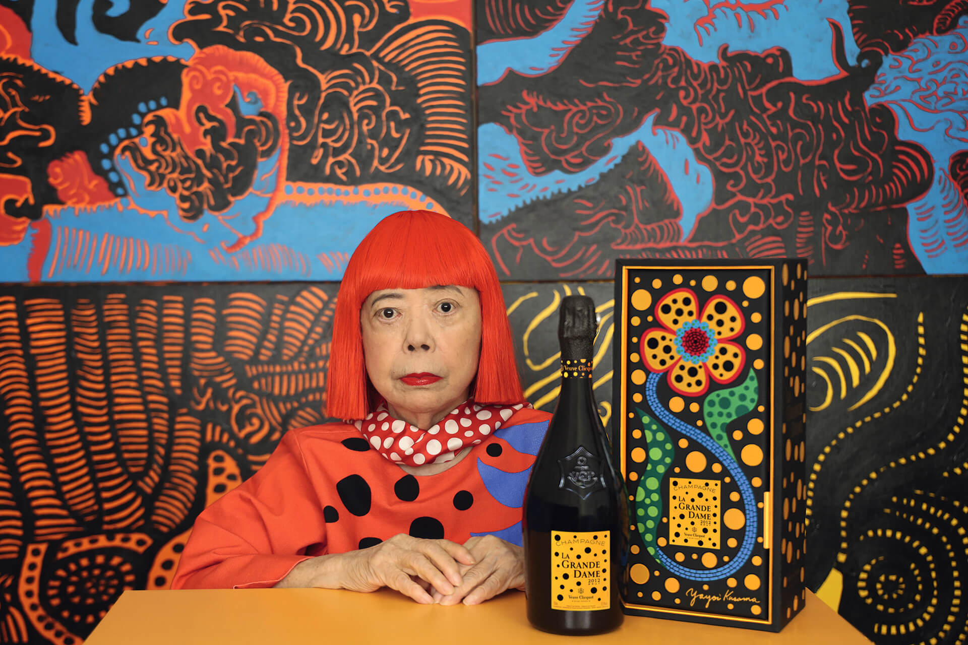 Artist in red wig and polka dot outfit next to a designed champagne bottle and box with a colorful abstract background.
