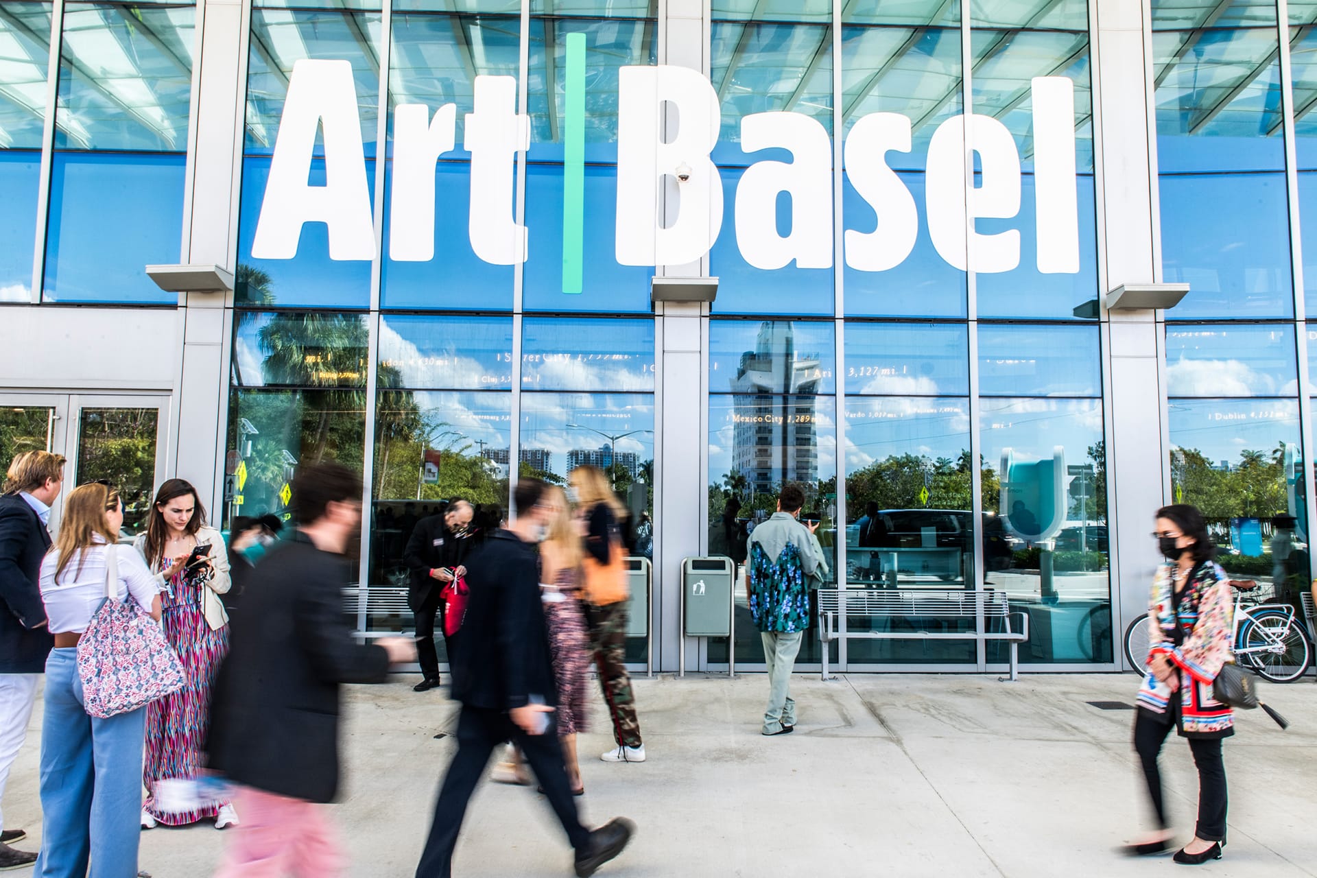 People walking in front of the Art Basel entrance with large glass windows displaying the event's name above them.