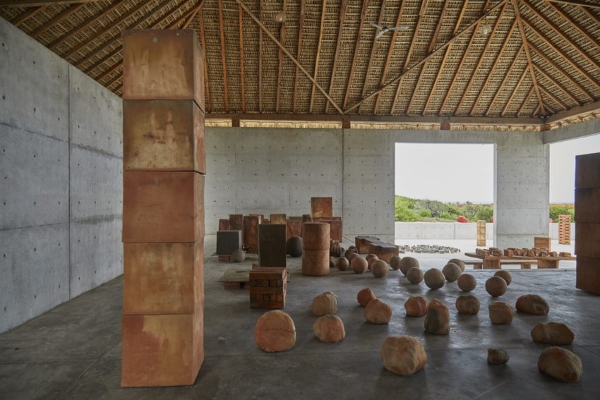 Indoor sculpture exhibition with stacked and scattered stone-like forms on a concrete floor under a wooden roof.