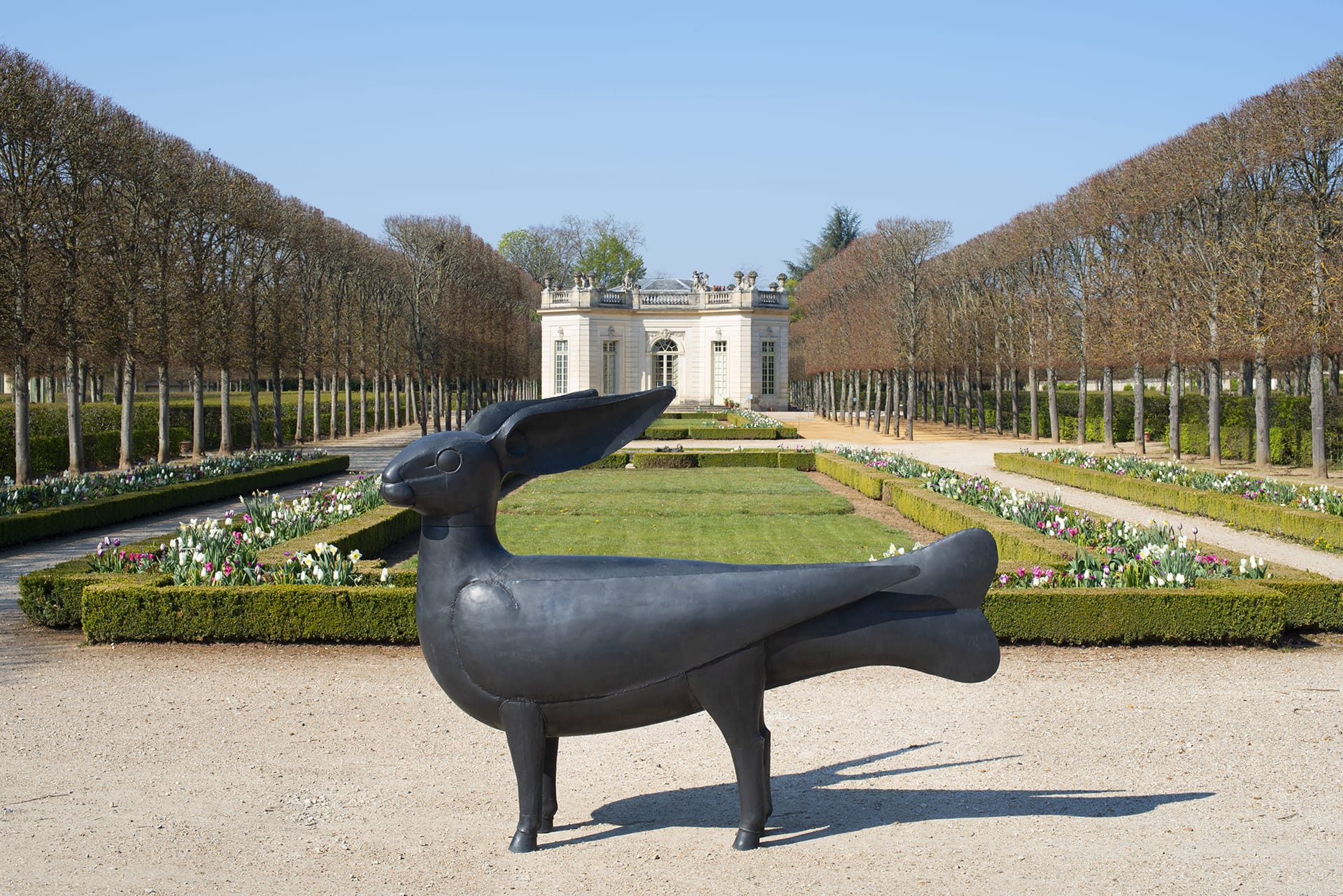 Sculpture of a hare with human legs in a formal garden with hedges and a small classical building in the background.