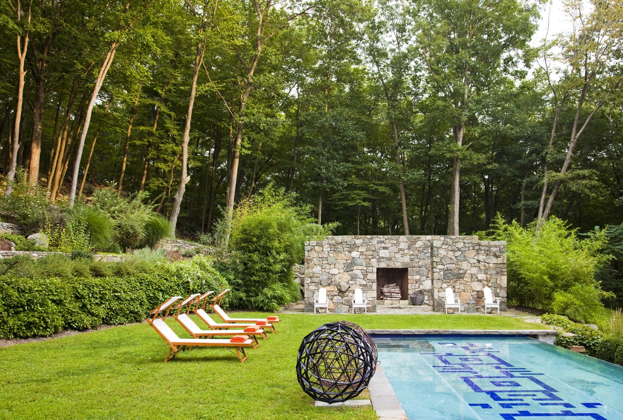 Contemporary backyard with a stone wall, swimming pool, lounge chairs, and lush green trees in the background.