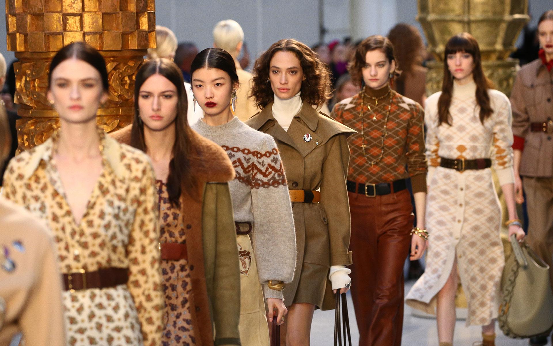 Fashion models walking down the runway showcasing a variety of stylish outfits with intricate designs and earthy tones.