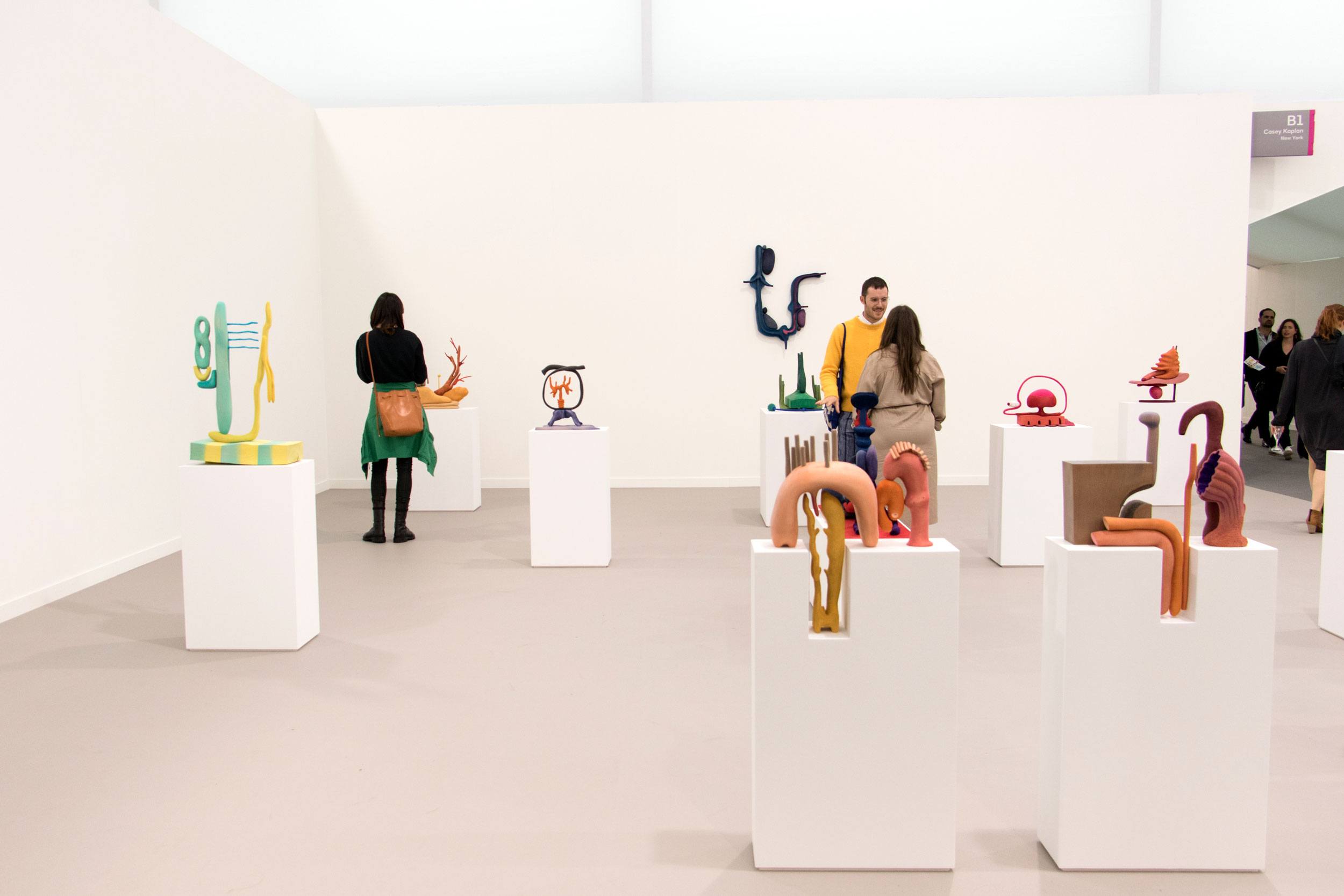 Art gallery visitors observing colorful abstract sculptures on white pedestals in a minimalist exhibition space.