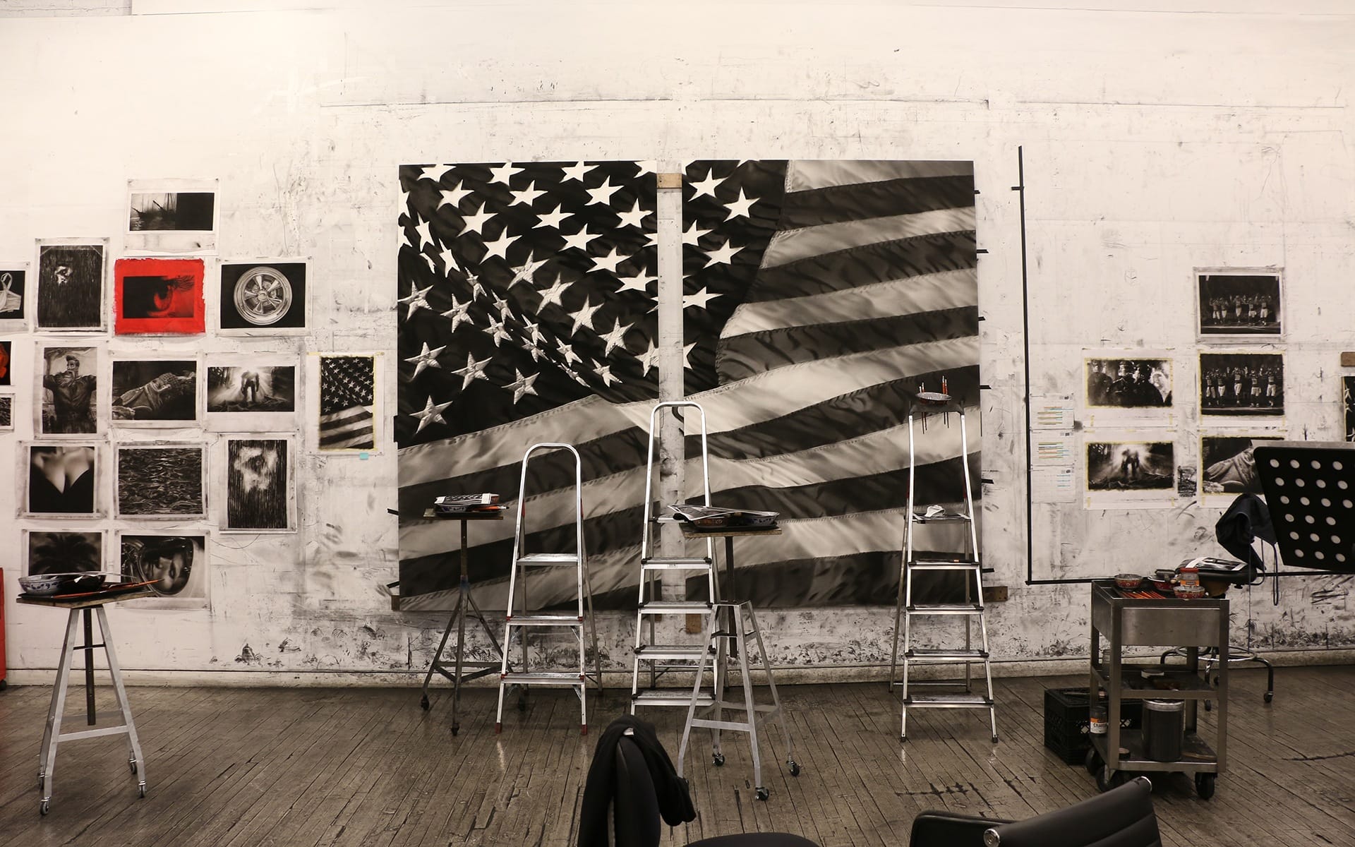 Art studio with American flag artwork, ladders, and tools in front of a wall displaying various black and white photos.