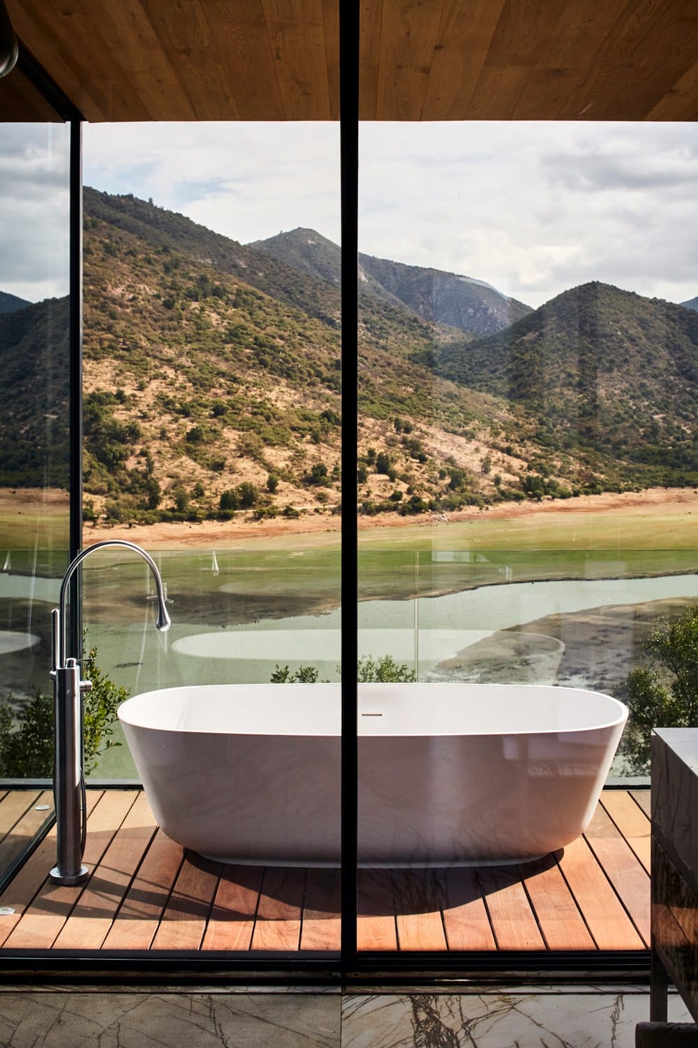 Modern bathtub with a scenic mountain view seen through large glass windows, set on a wooden floor.