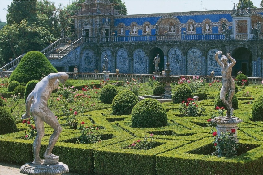 Lush garden with neatly trimmed hedges, statues, and a blue-tiled historical building in the background.
