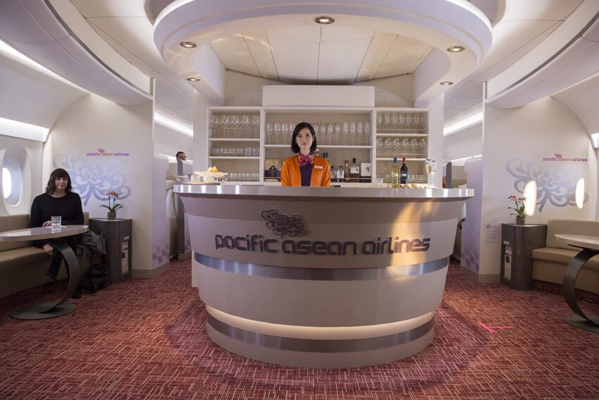 Flight attendant standing at a modern airline bar with passengers seated in a luxurious interior setting.