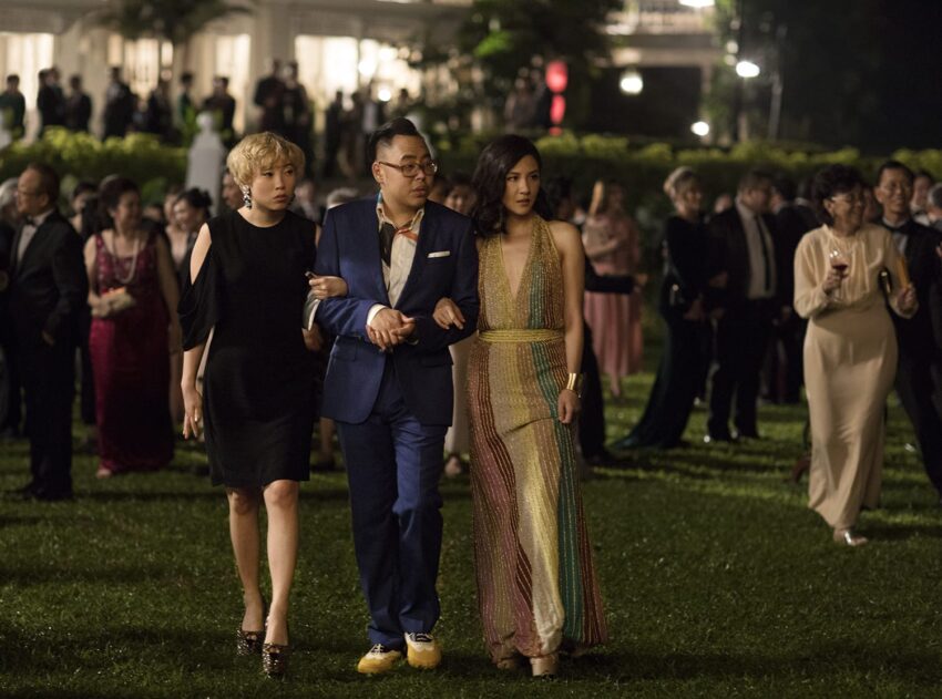 Three elegantly dressed people walking together at an outdoor event during the evening, surrounded by a crowd.