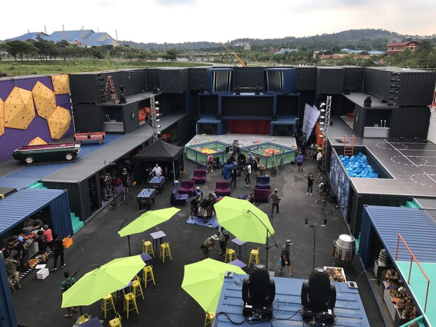 Outdoor set with colorful props and structures, people setting up, green umbrellas, and a scenic backdrop.