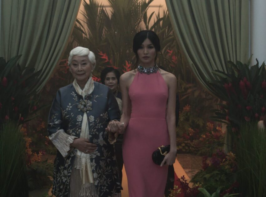 Two elegantly dressed women walking through a lush, exotic garden setting with vibrant green plants and red flowers