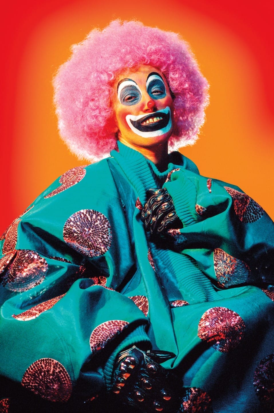A colorful clown with pink hair and bright costume, smiling against a vibrant orange-red background.