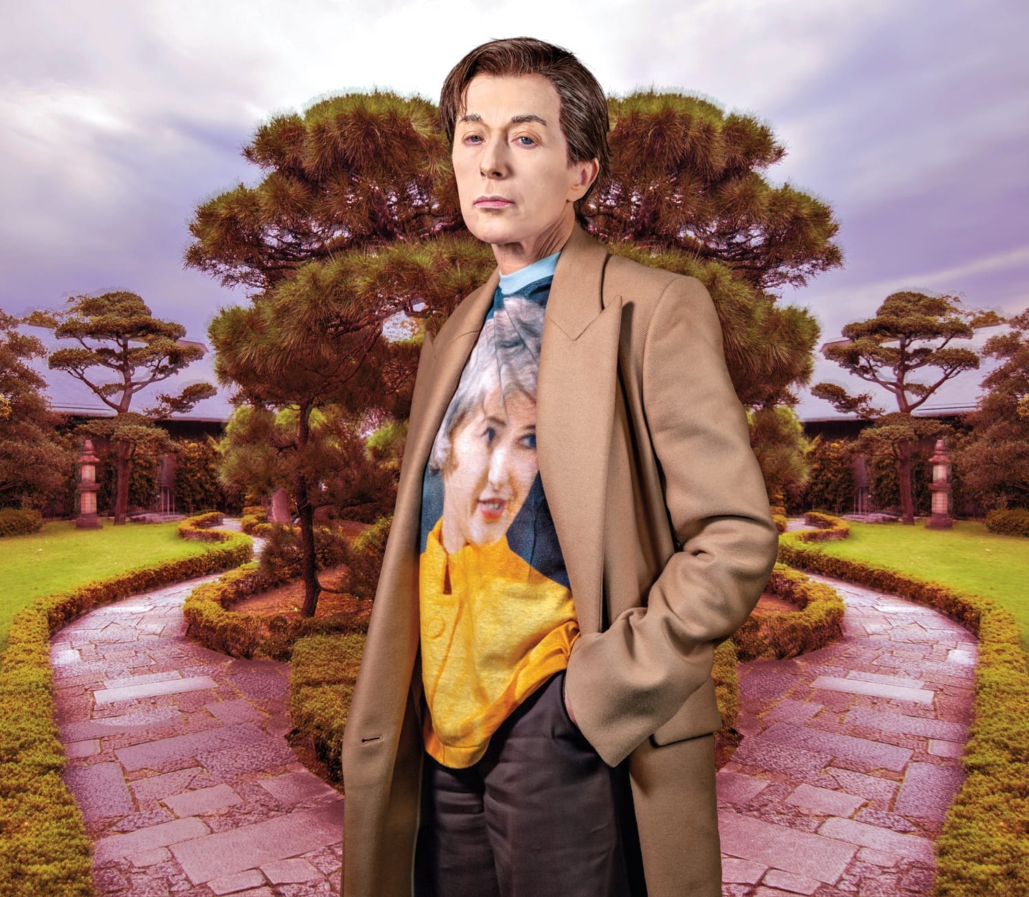 Person in a long coat stands on a garden path with trees in the background, wearing a colorful shirt.